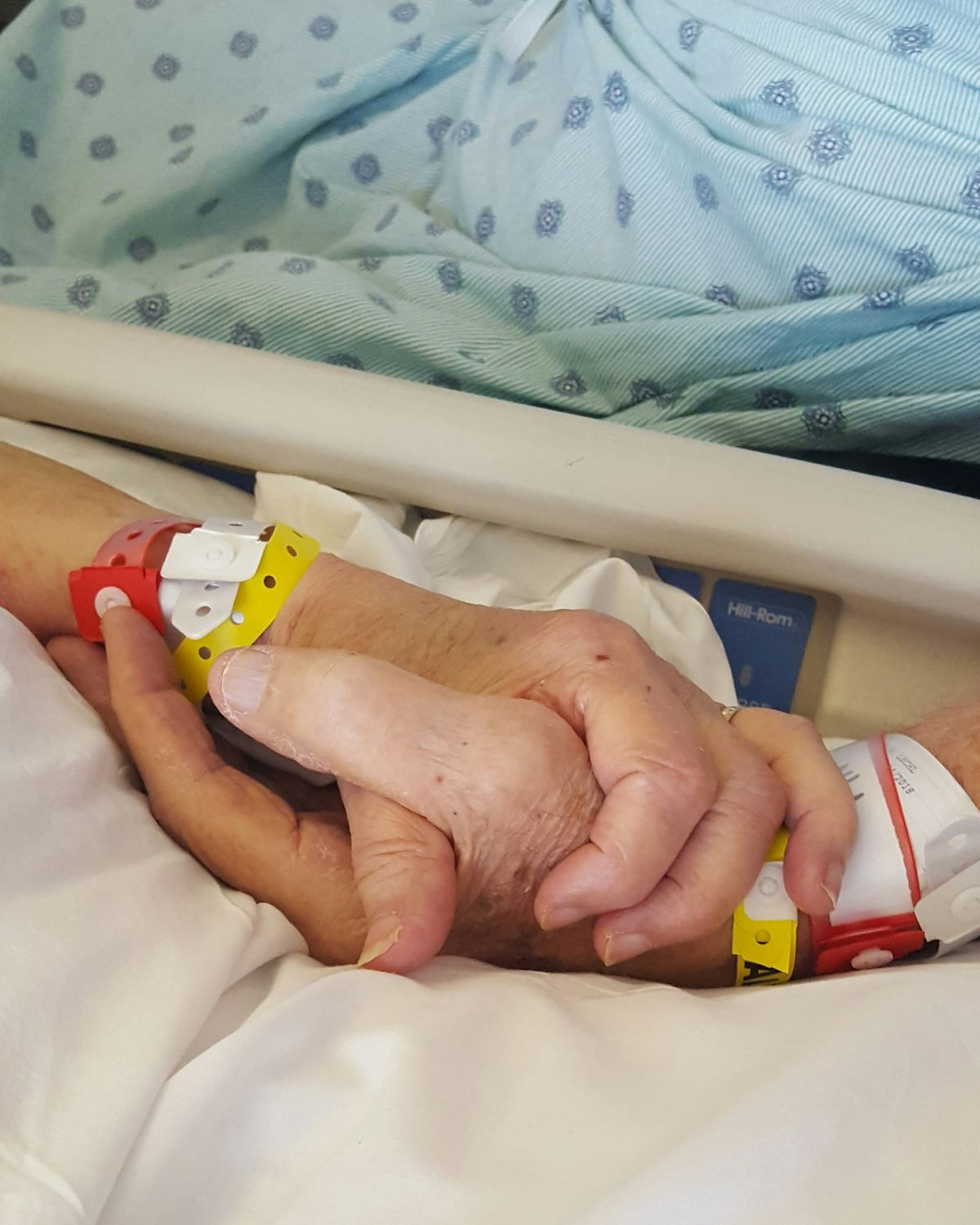 Mani intrecciate di due persone con i braccialetti del ricovero, accanto a un letto di degenza