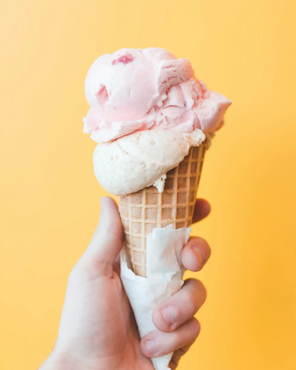 Mano che tiene un cono con due palline di gelato su fondo giallo